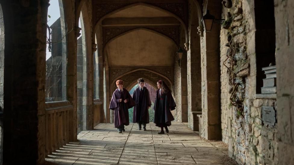Alastair Stout as Ron Weasley, Dominic McLaughlin as Harry Potter and Arabella Stanton as Hermione Granger walk the halls of Hogwarts in their wizarding robes in HBO Max's new Harry Potter series. 