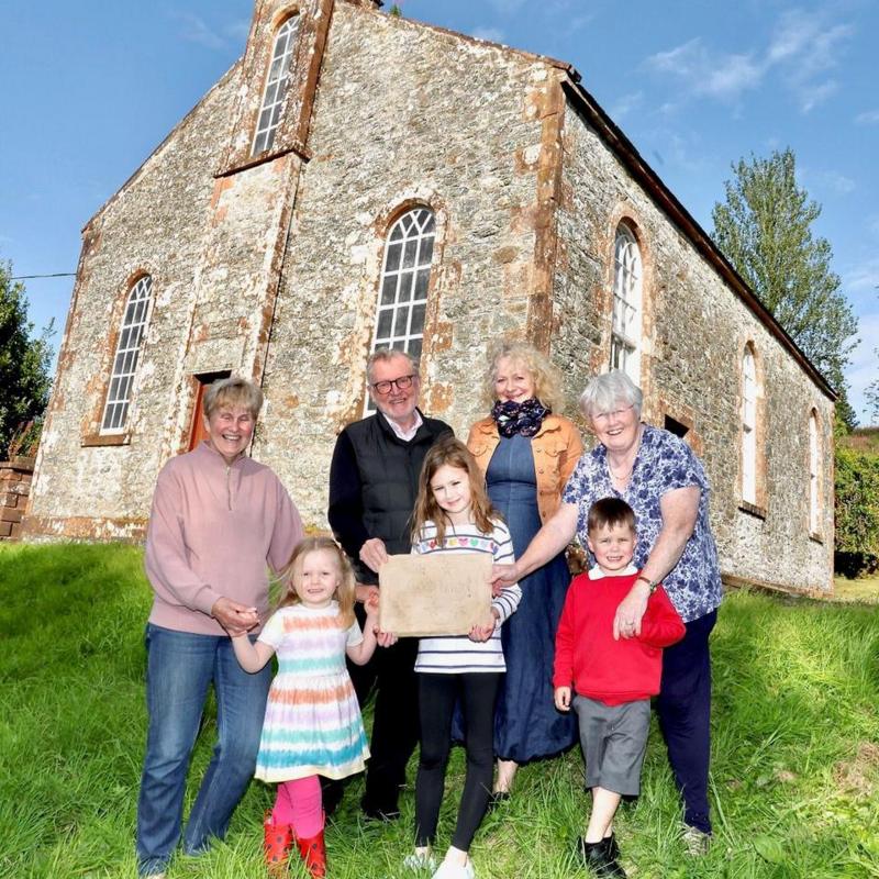 A group of four older people and three children stands in front of Wamphray Church on a sunny day