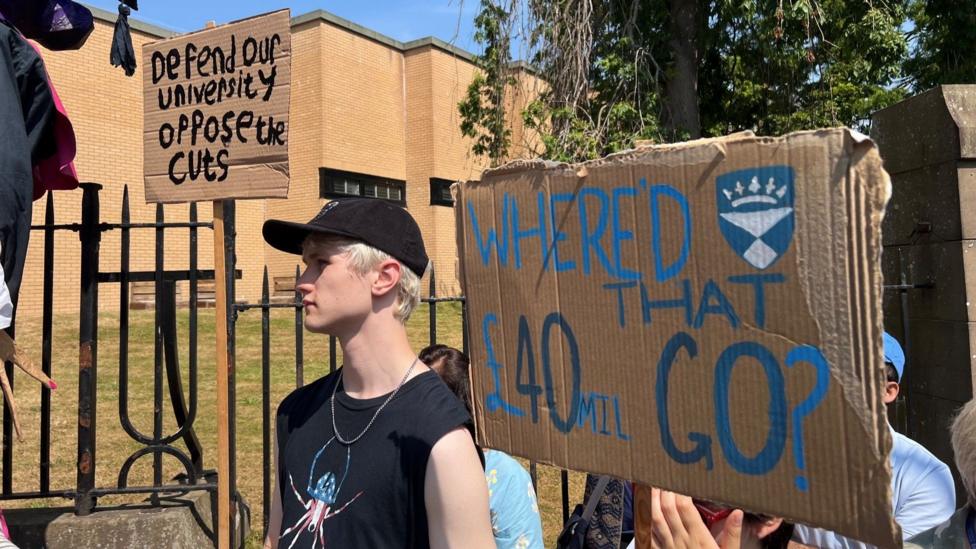 A protester wearing a black sleeveless T shirt and black baseball cap stands between two placards. One reads Defend Our University Oppose the Cuts and the other says Where'd That £40m go?