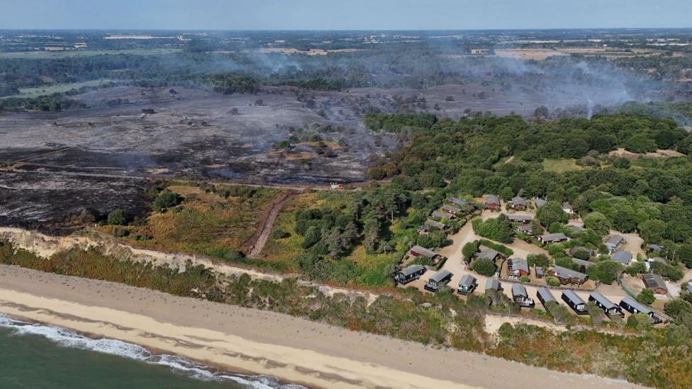 A drone image of a stretch of coastline. There is a holiday park with lodges. The land around the site has been scorched black from a wildfire. Smoke billows into the air from some of the land. 