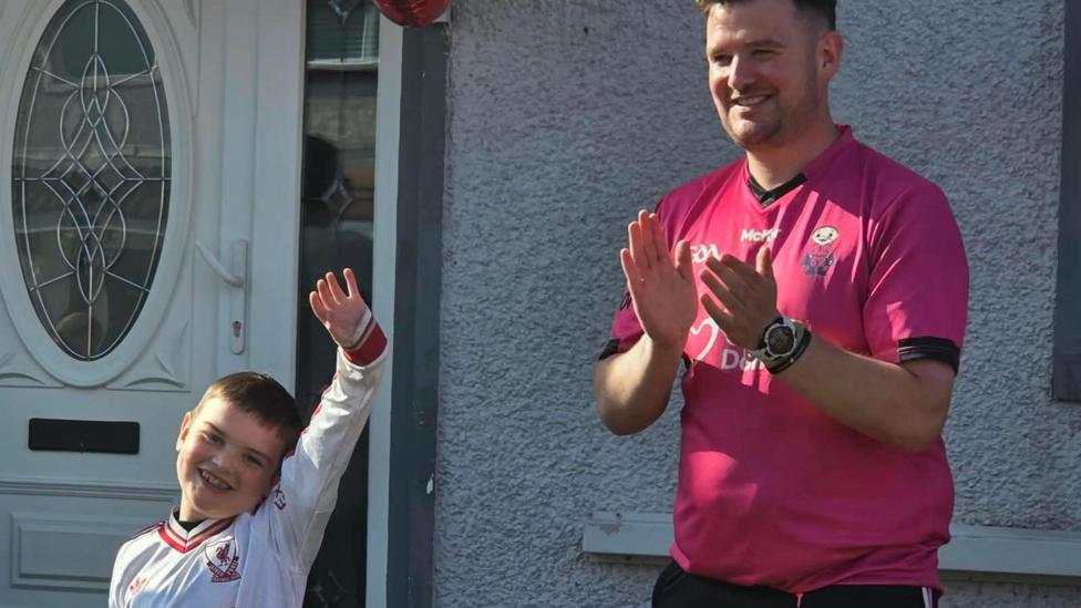 Dáithí is holding his hand up in the air. His dad Máirtín is clapping.
