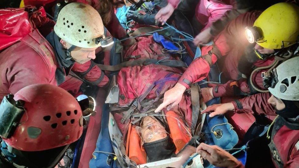 George Linnane is underground lying on a stretcher. He is surrounded by a rescue team who are wearing safety helmets with torches on the front of them.