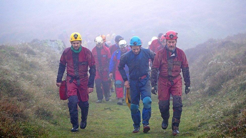 Several members of the rescue crew walking above ground following their efforts underground to help George Linnane. The air is misty. They are wearing safety helmets and workwear overalls.
