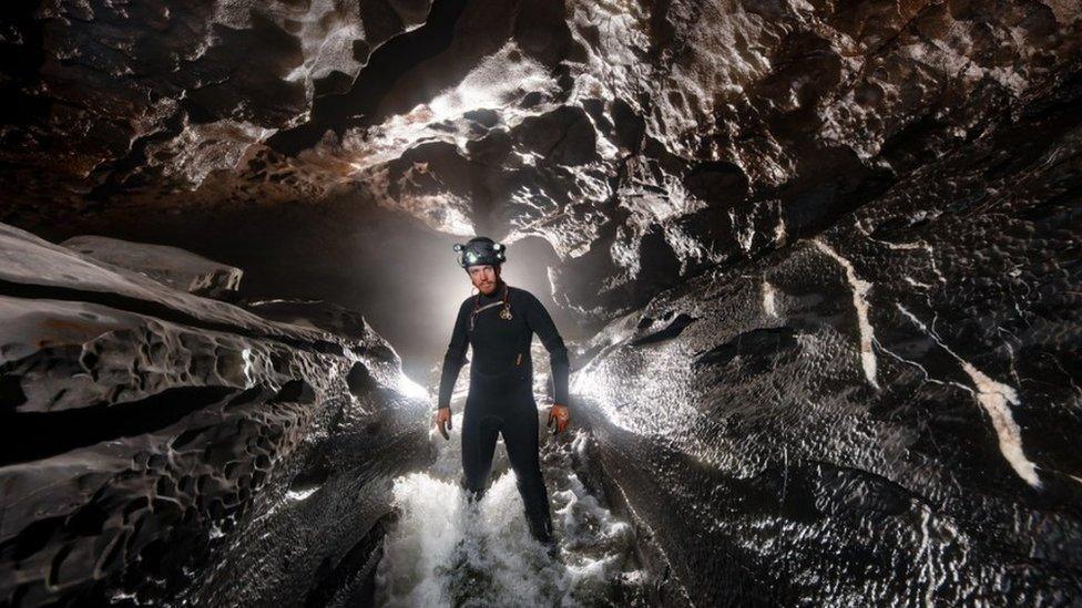 George Linnane is photographed underground in a cave. He is stood the centre of the image, standing in water. He is wearing a wet suit and a satfey helmet.