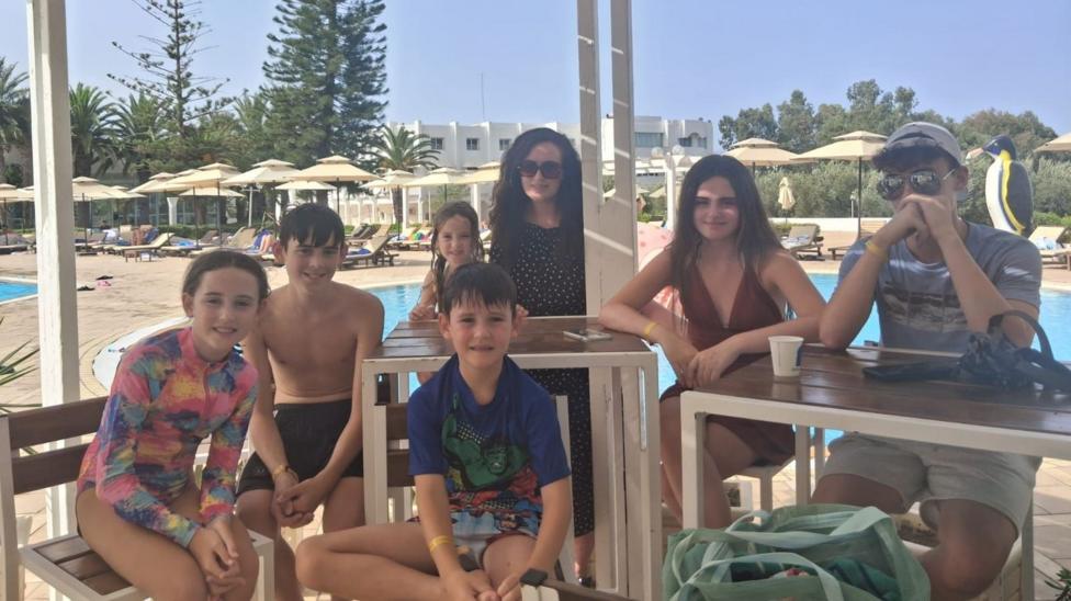 A mum and six children, of various ages, are sat at a poll side beach bar. all are wearing swimwear.