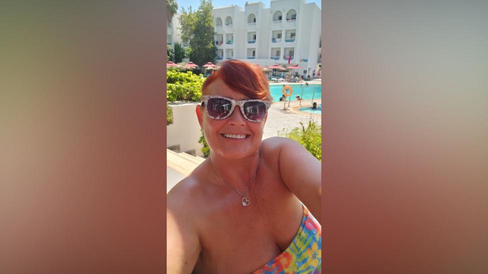 A woman is taking a selfie at a holiday resort. In the background is a swimming pool. She has ginger hair and is wearing a bikini. 