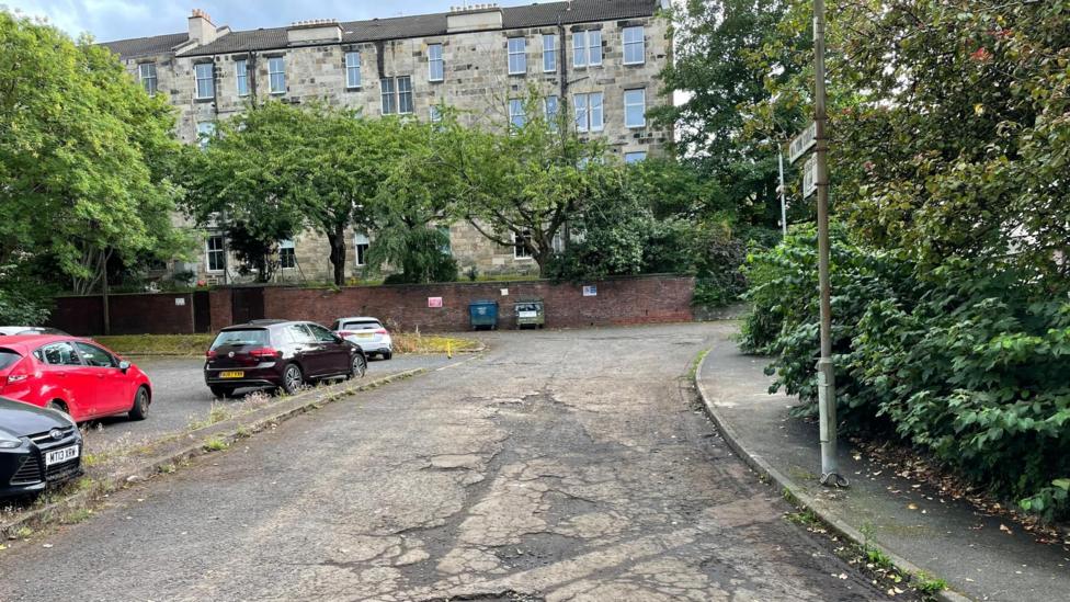Potholed road with cars parked next to it. There are flats, trees and a street sign and two bins against a wall in the distance