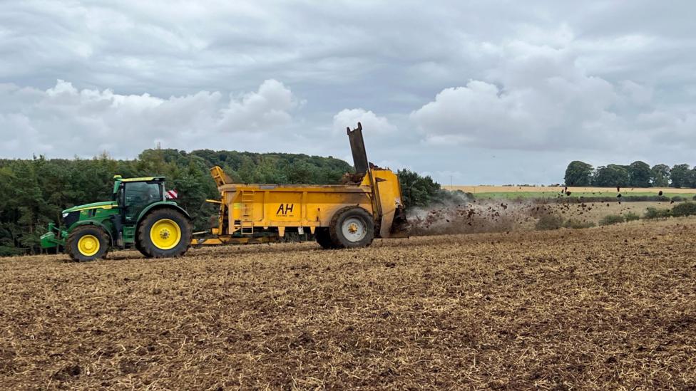 A green tractor is pulling a yellow trailer that is spreading muck on to a field of soil out the back of it.