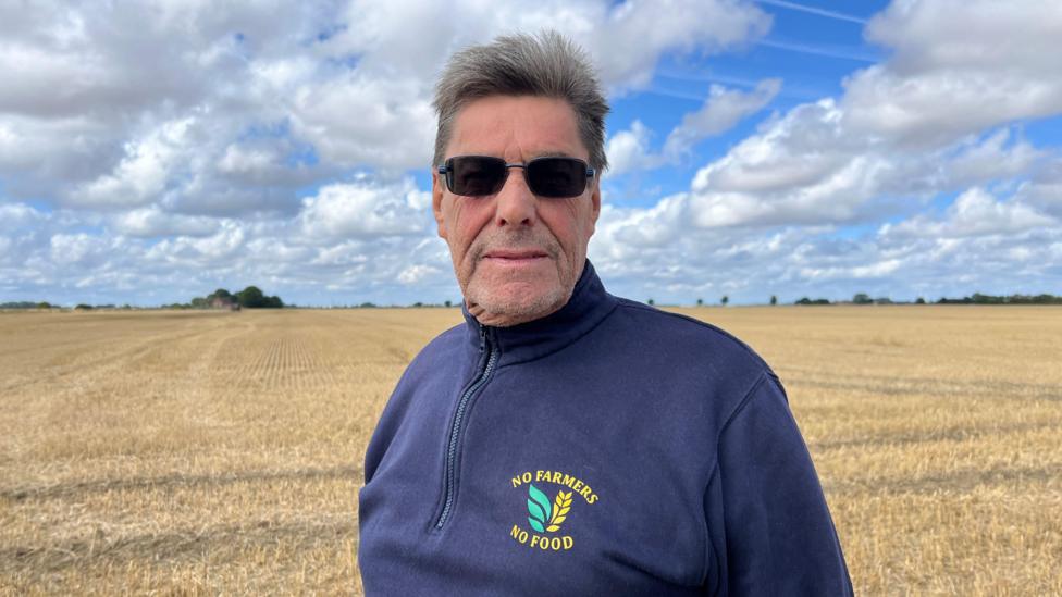 A man standing in a field and wearing a blue quarter-zip jumper. He has short grey hair and is wearing sunglasses as he looks directly into the camera.