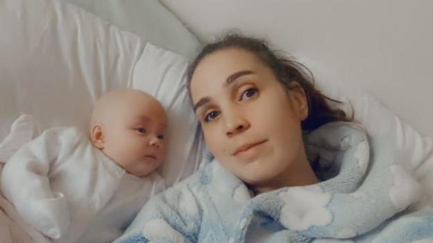 Natalie is lying on a bed beside a baby dressed in a white outfit. The person is wearing a light blue fleece robe with a cloud pattern, and both are positioned against white pillows and bedding.