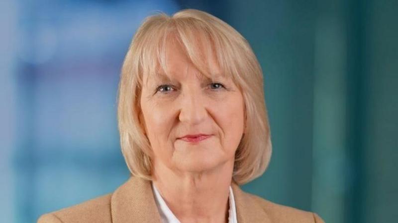 A picture of Jacqui Durkin, taken from the shoulders up. She has shoulder length blonde hair with a fringe and is wearing a white shirt and beige blazer. There is a blurred blue background behind her. 