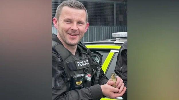 PC Clough in his police uniform including a thick black stab vest smiles as he holds a small yellow duckling. 