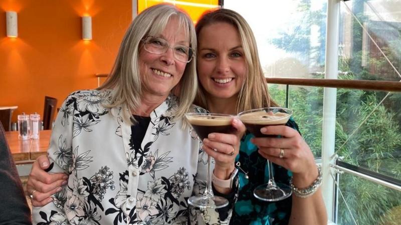 A woman in her 60s with shoulder length blonde hair and frameless glasses sits smiling next to a younger woman who is also smiling and who also has blonde hair to her shoulders. They are both holding espresso martini cocktails and they are sitting in a restaurant with orange walls to the left and a large window to the right, where bamboo can be seen growing.