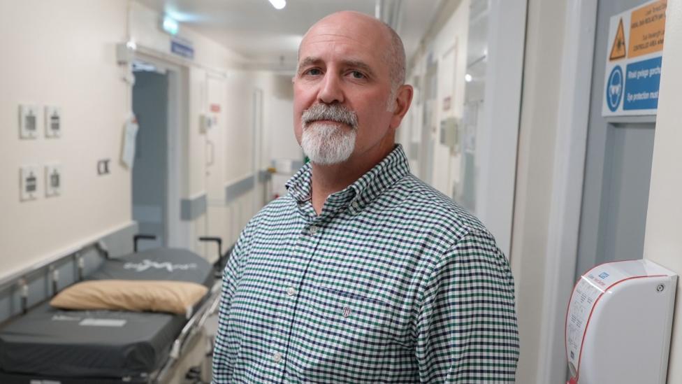 A man stands in a hospital corridor with a trolley to his left and a closed door and hand sanitiser unit mounted on the wall to his right. He is bald with a short white goatee beard and is wearing a green, white and black gingham shirt.