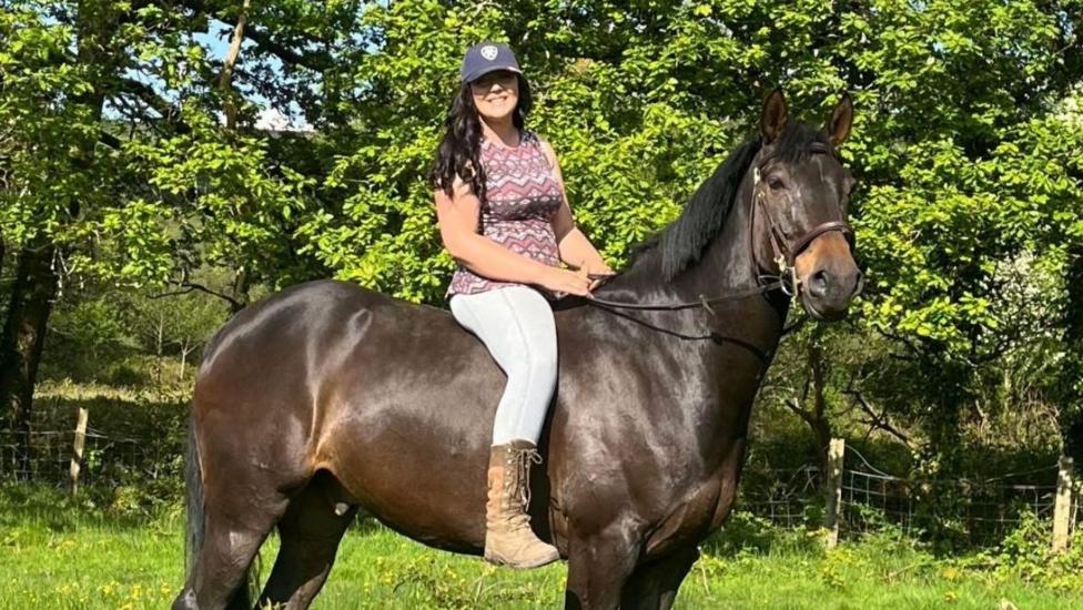 A woman, Tasha Lee Hopkins, sits without a saddle on a large, dark brown horse in a field and smiles into the camera. She is wearing a cap, patterned top, light blue leggings and tan lace-up boots. Her long brown hair is down.