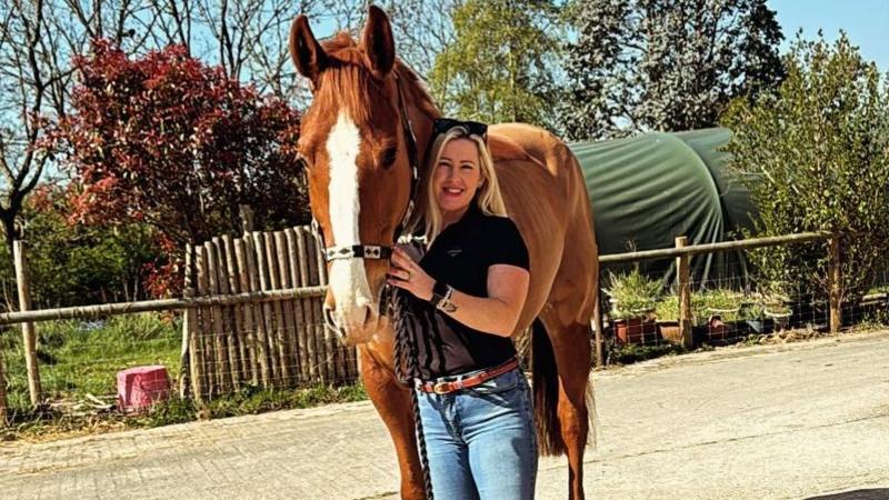 A blonde white woman with black sunglasses over her head wearing a black shirt and blue jeans is standing next to and holding a brown horse with a white patch on its nose.