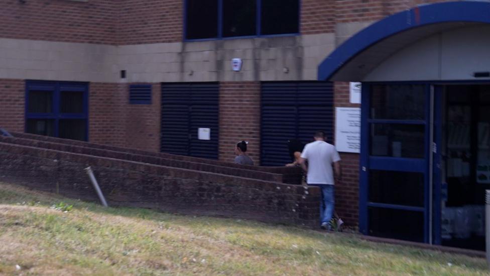 A blurred image of a woman, Tasha Lee Hopkins, walking out of a court building with her back to the camera. She is wearing a black and white striped top with her dark hair, which has a blonde streak in the front, tied up in a bun on top of her head. A man and a woman are walking behind her.