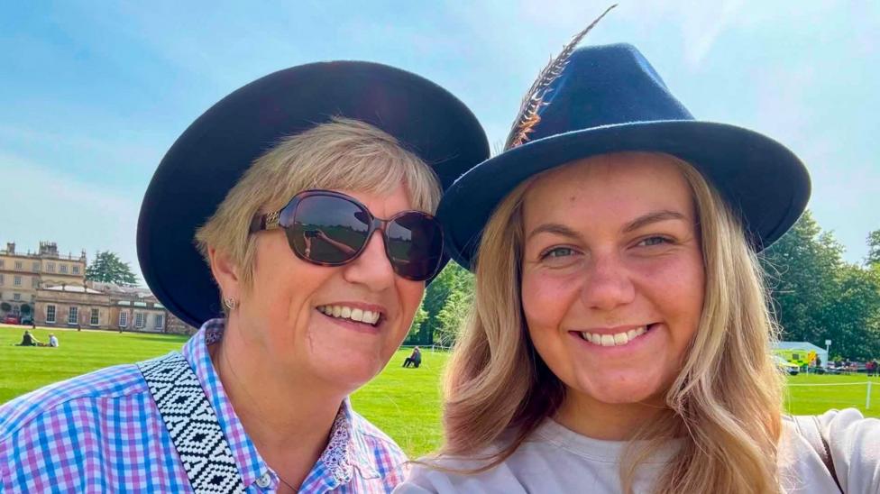 A picture of two blonde women side by side, the woman on the left has short hair with a dark hat on. She is also wearing a blue and pink check shirt and dark sunglasses. The woman on the right has long hair, with a navy blue hat on and is wearing a grey top.