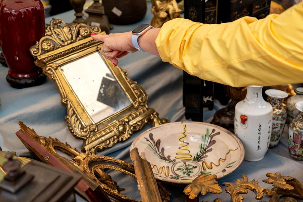 A shopper in a yellow jacket points at an ornate gilt mirror among vintage ceramics and decorative frames on a flea market stall.