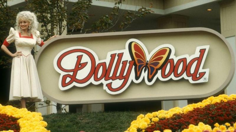 A smiling Dolly Parton poses in front of the Dollywood sign in 1988. She is wearing a long white dress with a red trim around the neckline and arms.