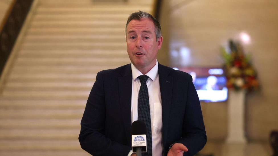 a man in a black suit blazer with a skinny black tie over a white shirt, speaking at a Northern Ireland Assembly - branded microphone in a room that is recognisable as Stormont's great hall