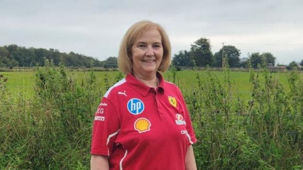 Margaret Peoples is wearing a red ferrari shirt and is standing in a grassy area. She has long blonde hair. 