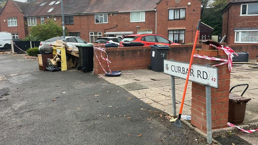 A street sign reading Curbar Road with houses and cars behind it. Red and white tape is wrapped around walls and there is debris in the street.