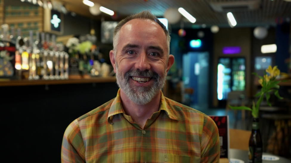 Man in the foreground wearing a yellow and green checked shirt, seated indoors and facing the camera. The setting is a bar or pub, with a row of beer taps and bottles visible behind the person on the left. Tables and chairs are positioned in the background, and a small vase containing yellow flowers sits on a table to the right. Overhead ceiling lights and hanging decorations are visible throughout the venue. No bags, bins or markers are visible.