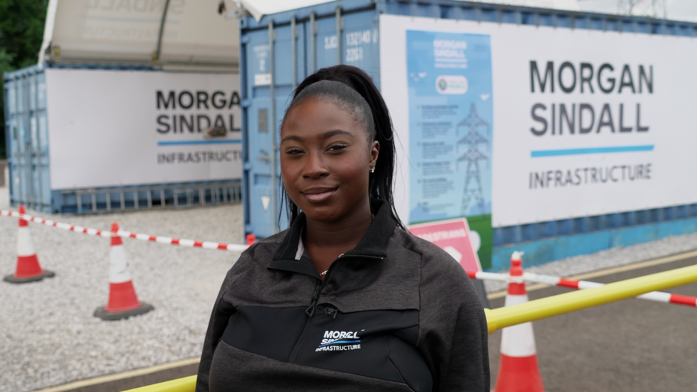 A young woman in a black Morgan Sindall Infrastructure zip-up jacket standing in the foreground at a construction or infrastructure site. Behind the person are two blue site cabins or container units displaying large Morgan Sindall Infrastructure signs. Red-and-white traffic cones and barrier tape mark off a gravel area, with a yellow safety barrier running across the lower right of the image. Additional site signage is attached to one container, and electricity pylons are visible in the background.