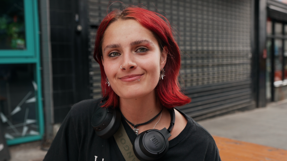 young woman with short red hair in the foreground, wearing a black T-shirt, black over-ear headphones around the neck, a cross-body strap, a nose ring and earrings. The person is facing the camera on a pavement outside a building. Behind the person is a dark tiled wall and metal security shutter, with a turquoise-framed doorway visible on the left and part of a street scene on the right. No bags, bins or markers are visible.