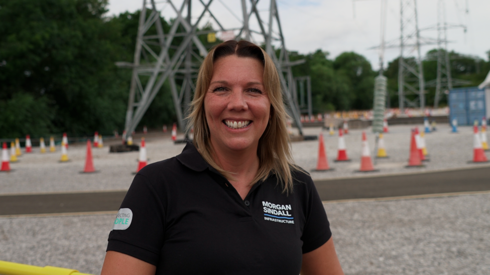 woman in the foreground wearing a black polo shirt with a “Morgan Sindall Infrastructure” logo, standing at an outdoor electrical infrastructure site and facing the camera. Behind the person are large steel electricity transmission structures, overhead power lines, and a gravel-covered work area. Numerous orange-and-white traffic cones mark pathways and restricted areas across the site. Trees line the background, and a blue site container is visible on the right. No bags or bins are visible.