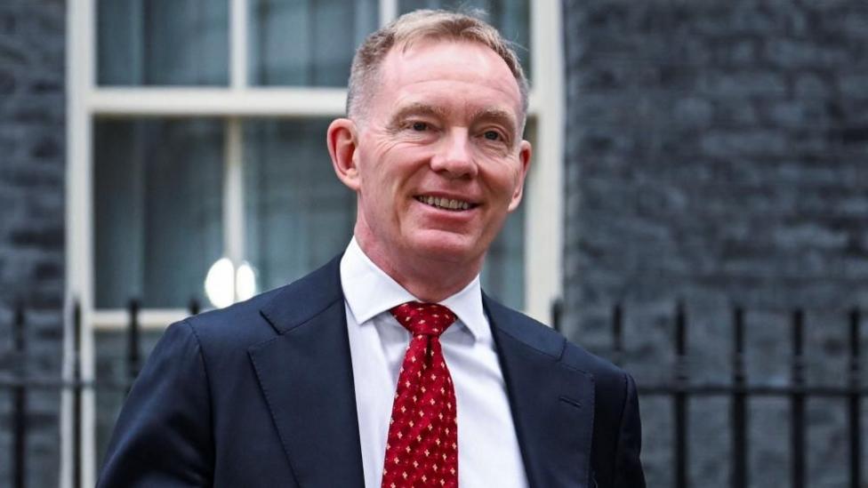  Sir Chris Bryant outside Downing Street. He is smiling. He's wearing a black suit with a white shirt and red tie.