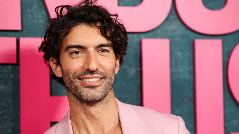 Justin Baldoni in a pink suit posing in front of a grey backdrop with large pink letters at a film premiere