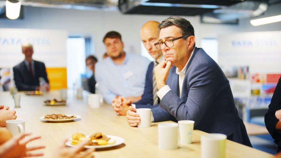 Andy Burnham, wearing a blue jacket and white shirt, sits at a table surrounded by people while he listens to someone speak. He has a serious look on his face.