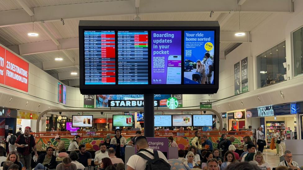An airport departures lounge that is filled with people sitting in chairs and at tables. A sign detailing flight information shows a lot of red squares for cancellations and delays.