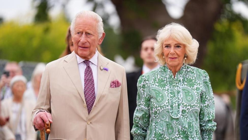 King Charles and Queen Camilla standing side by side on Sandringham Estate. The King wears a light beige suit and has his right hand resting on a wooden shepherd's crook. The Queen wears a green, paisley print dress. A crowd is blurred in the background.
