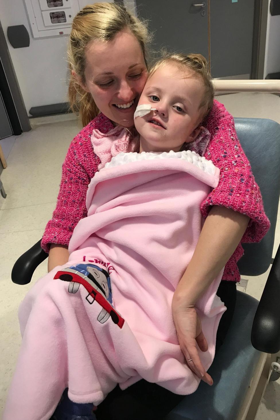 A young blonde girl with a feeding tube. She is wrapped in a pink blanket and is sat on the knee of woman in a pink jumper who is looking down on her with a smile. They are sat on a chair in a hospital.