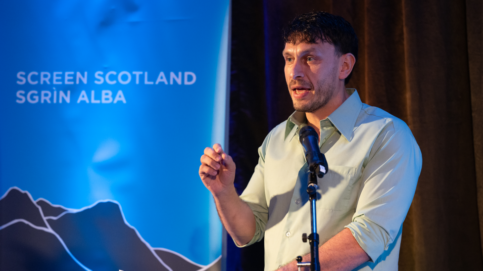 Richard Gadd is speaking at a microphone. He has short, dark hair and a short beard and he is wearing a grey shirt. His mouth is open mid speech and his arm is raised. He is standing beside a blue screen that says Screen Scotland and depicts mountains