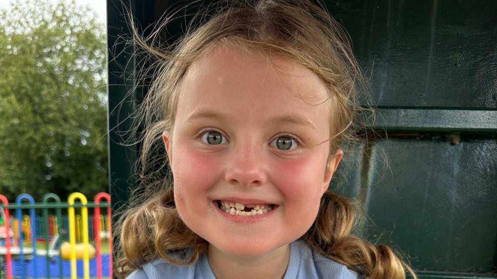 A young girl with curly hair in two bobbles. She is smiling and looking at the camera.