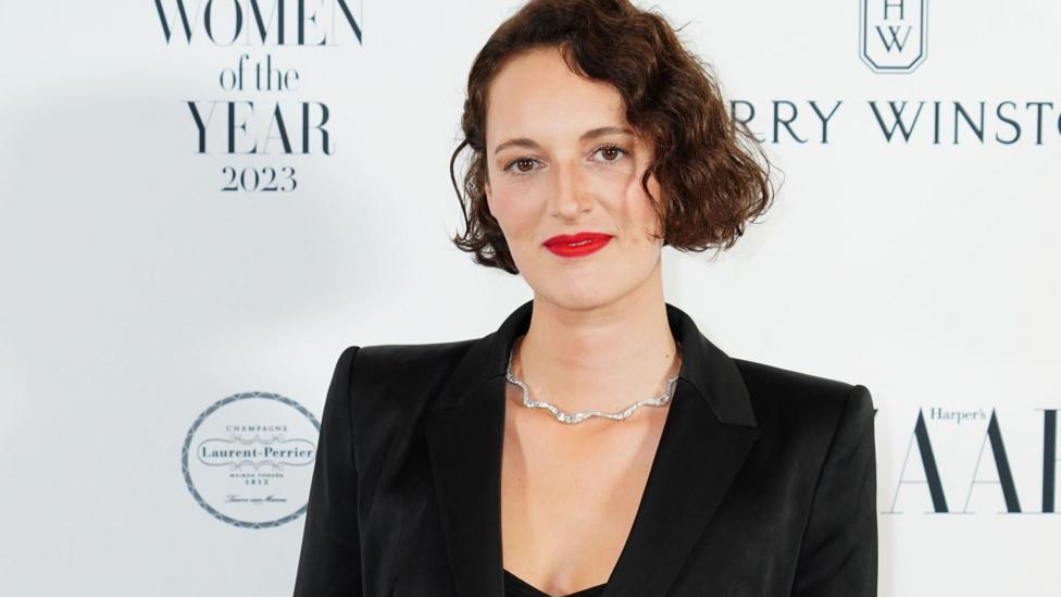 Phoebe Waller-Bridge, a tall woman in a smart tailored black jacket, sleek wavy bob and red lipstick. She is posing for a photo at some kind of red carpet event.