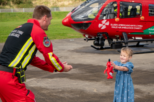 A young girl in a blue dress holding a red stuffed animal. She is reaching out to a paramedic in red and black Air Ambulance uniform who also has his hands reached out towards her. Behind them is a red air ambulance helicopter.