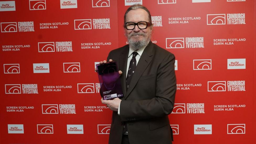 Sir Gary Oldman poses with his Global Icon award, a purple glass rectangle. Gary wears a chocolate brown suit, his hair is pulled back and he has a substantial beard in salt and pepper tones.