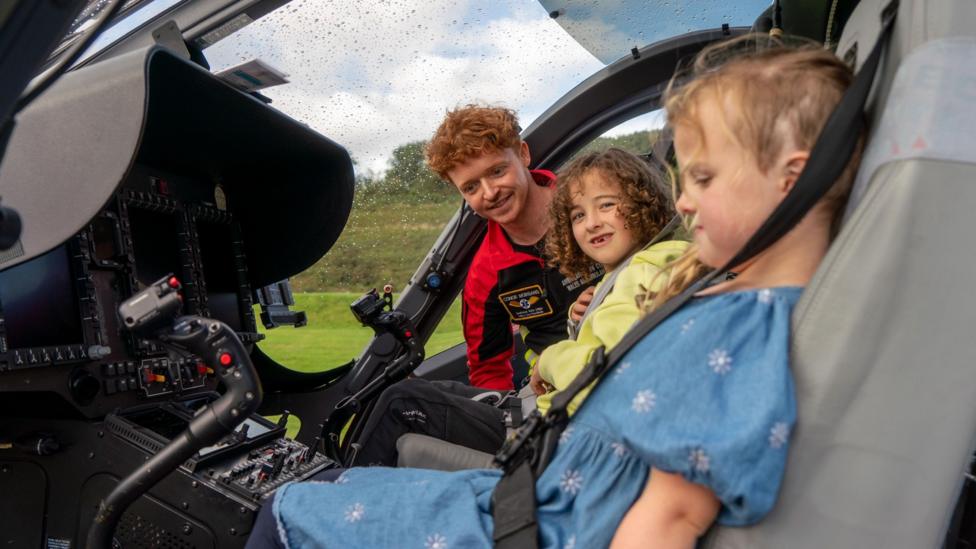 A young blonde girl and young brunette boy sat in a helicopter which is on land. Stood at the door beside them is a young man with ginger hair.