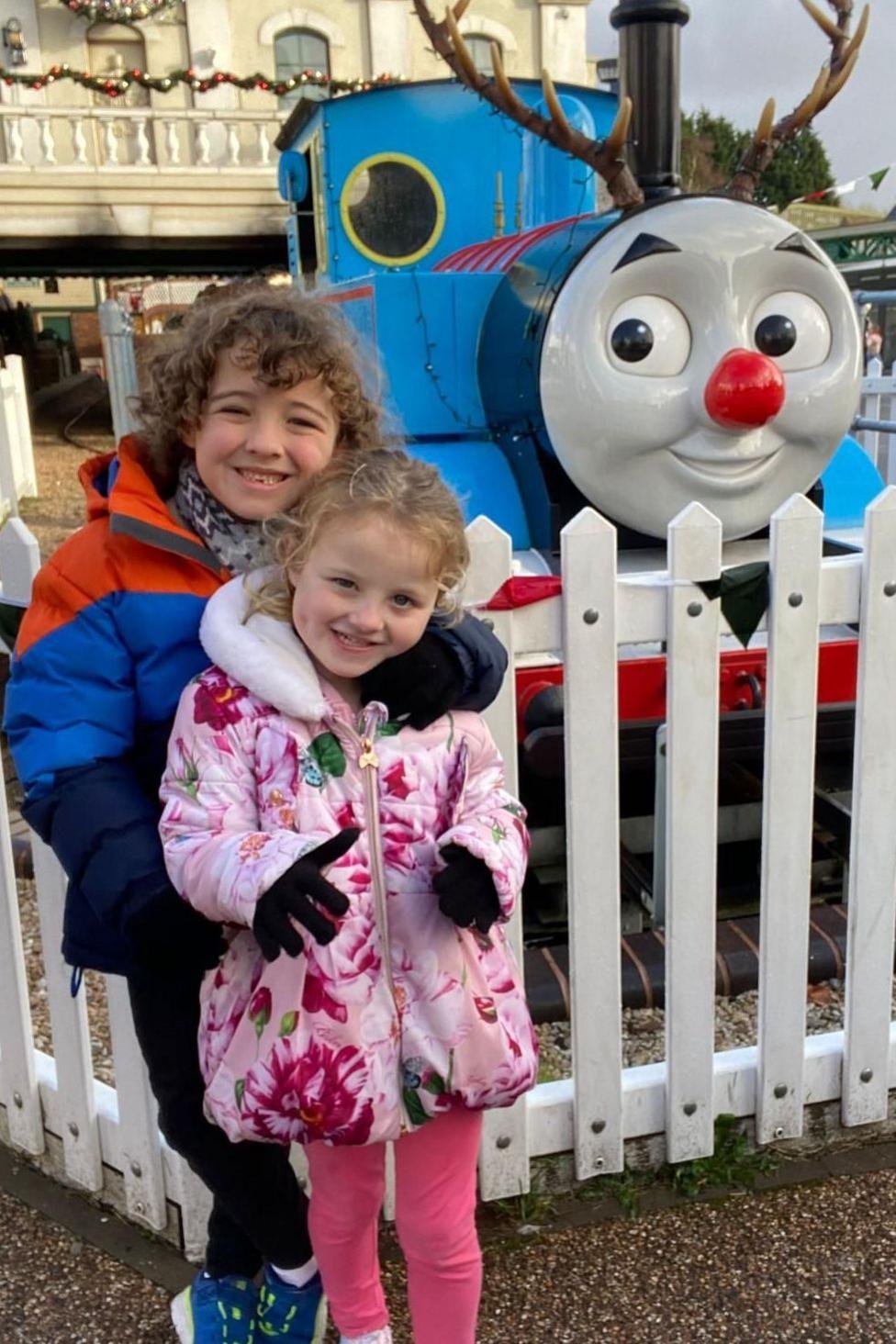 A young brunette boy with his arm around a young blonde girl. They are dressed in warm winter coats, scarves and gloves and stood with a large Thomas the Tank Engine model behind them. They are both looking at the camera and smiling.