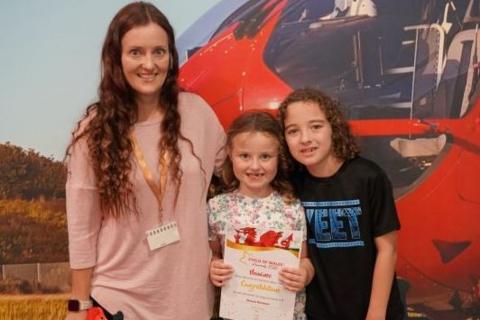 A woman with long, curly dark hair wearing a pink jumper. She has her arm around two children - a young blonde girl and a young brunette boy. The girl is holding a certificate and they are all looking at the camera and smiling. Behind them is a poster with a picture of a red helicopter.