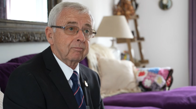 An older man wearing glasses, a dark suit and striped tie sits on a purple sofa in a living room, looking towards the camera.