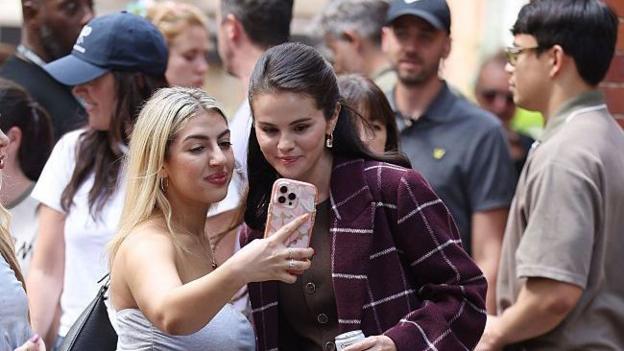 Selena Gomez in a red checked coat next to a fan who is taking a selfie of them both together.