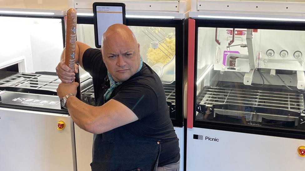 Taking the stance of a baseball batter, Lee Kindell holds a roll of pizza dough as if it's a bat and makes a mock-serious game face. 