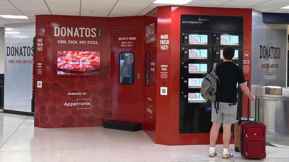 The dark red Donatos fresh pizza vending machine is about the size of a van. A customer is looking at its screen.