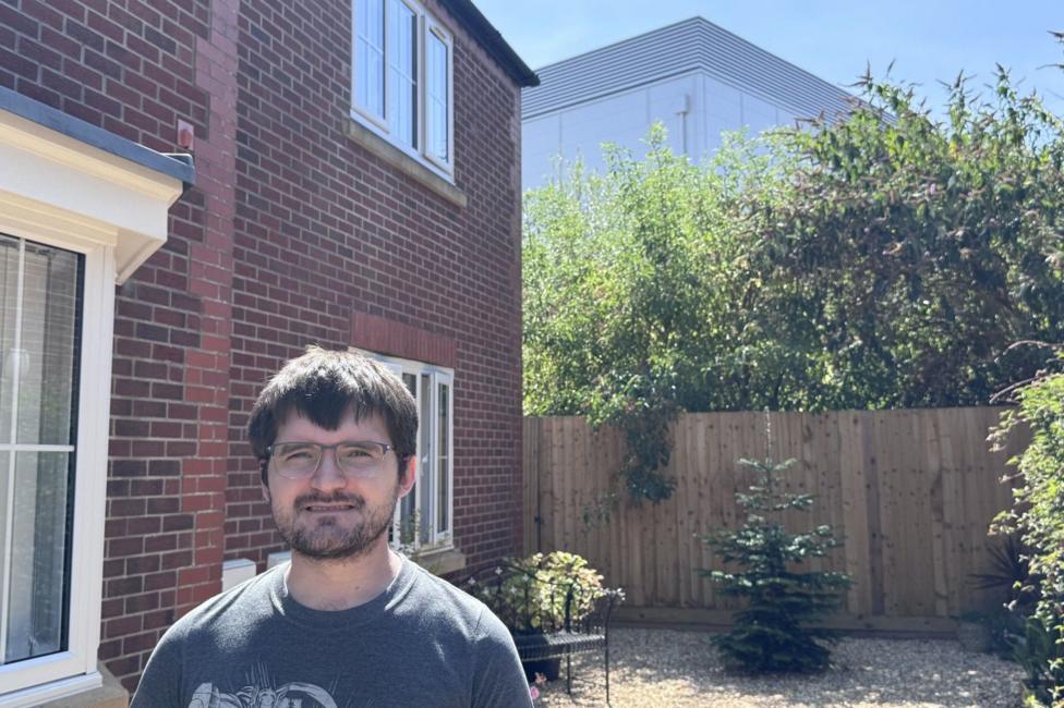 A man with short brown hair stands in his back garden. Behind him beyond the fence is a data centre which looks like a tall warehouse